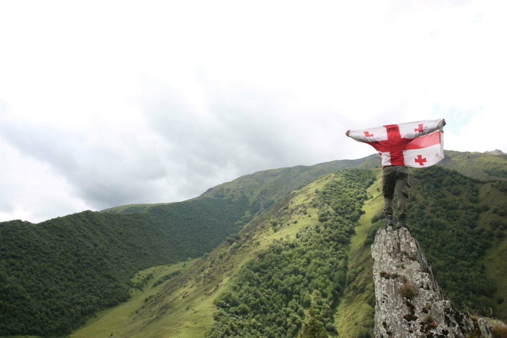 Człowiek stoi z flagą Gruzji na szczycie góry przed tłem panoramy leśnej
