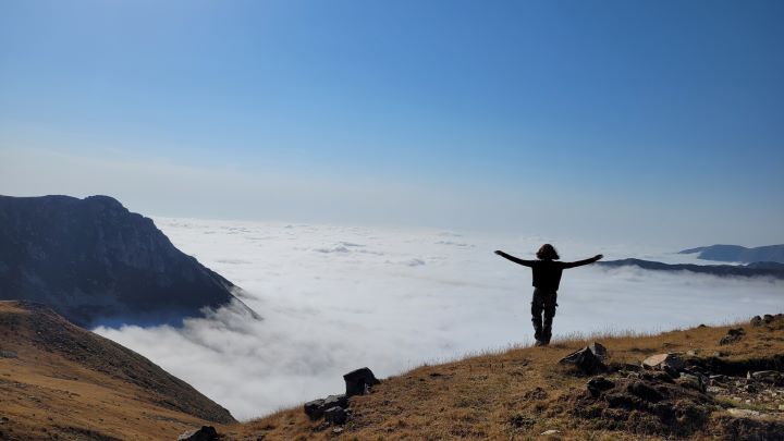 Kobieta stojąca na szczycie góry z szeroko otwartymi ramionami nad doliną wypełnioną chmurami