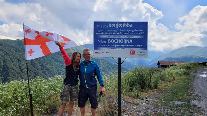 Kobieta i mężczyzna w ubraniach outdoor stoją trzymając flagę Gruzji przed tablicą, która oznacza "Village Bochorna - Welcome to the highest settlement in Europe" w języku gruzińskim i angielskim, na tle zielone góry i doliny