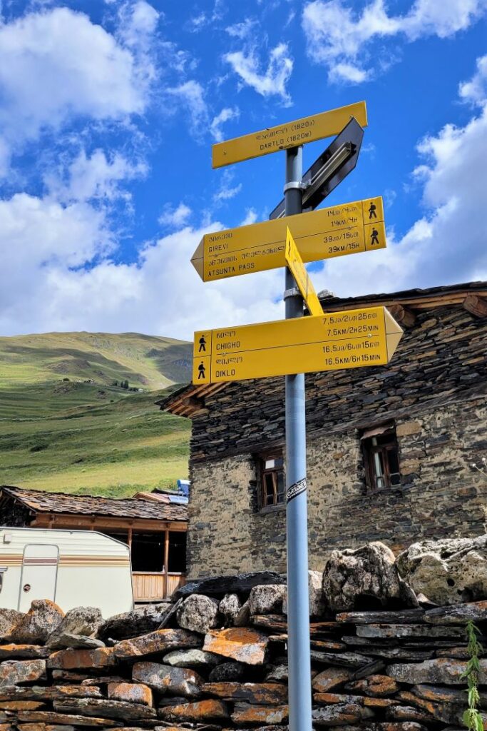 Drogowskaz żółty dla wędrujących w Tuszetii, Gruzja, pokazując drogi do Atsunta Pass, Chigho i Dartlo; na tle karawan i dom z kamienia